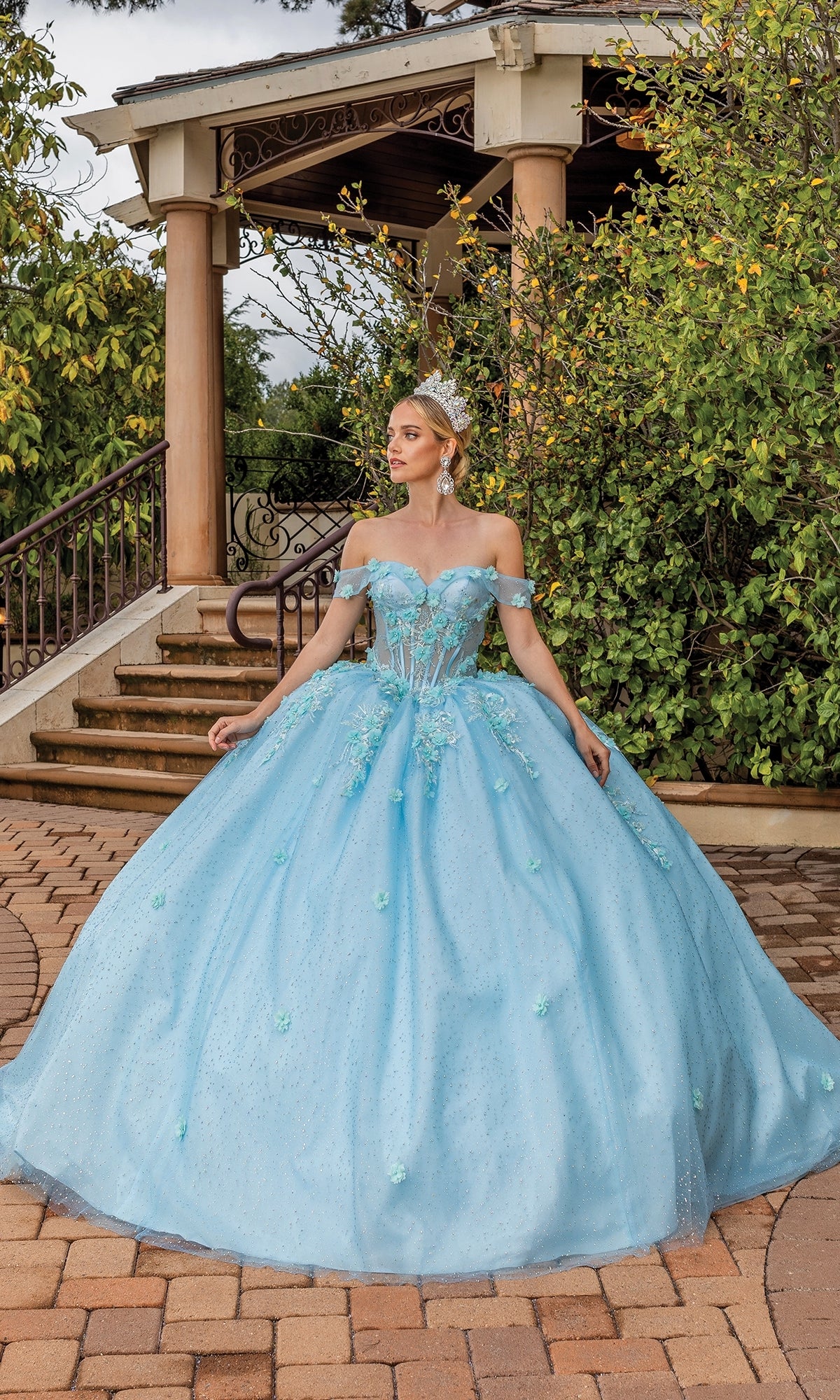 Wearing the Quinceanera Dress 1867 By Dancing Queen, a woman stands on a stone path near a gazebo, surrounded by lush greenery.